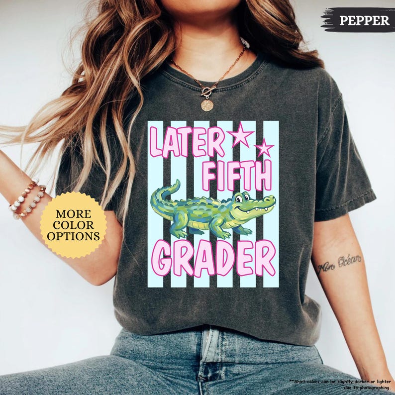 May include: A dark gray t-shirt with a graphic that says "Later Fifth Grader." The design features a cartoon alligator in front of light blue stripes and pink text. The shirt also has the word "PEPPER" printed on the top right.