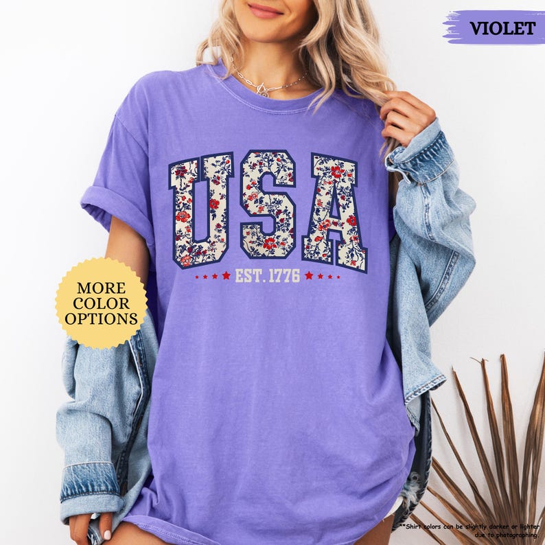 May include: A purple t-shirt with a floral print of the word "USA" and the text "EST. 1776".