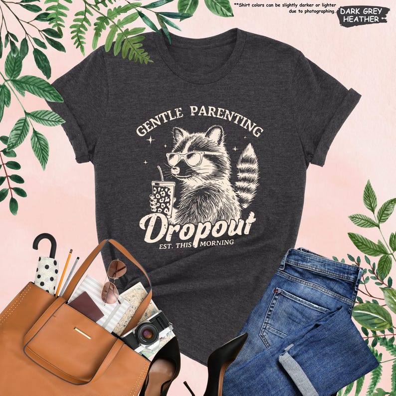 May include: Dark grey heather t-shirt with a raccoon wearing sunglasses and holding a drink. The shirt features the text "GENTLE PARENTING Dropout EST. THIS MORNING". The shirt is paired with blue jeans and a brown tote bag.