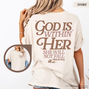 May include: Ivory-colored t-shirt with the text "GOD IS WITHIN HER SHE WILL NOT FALL PSALM 46:5" in brown and mauve lettering. The shirt has a small graphic on the front and is a casual style.