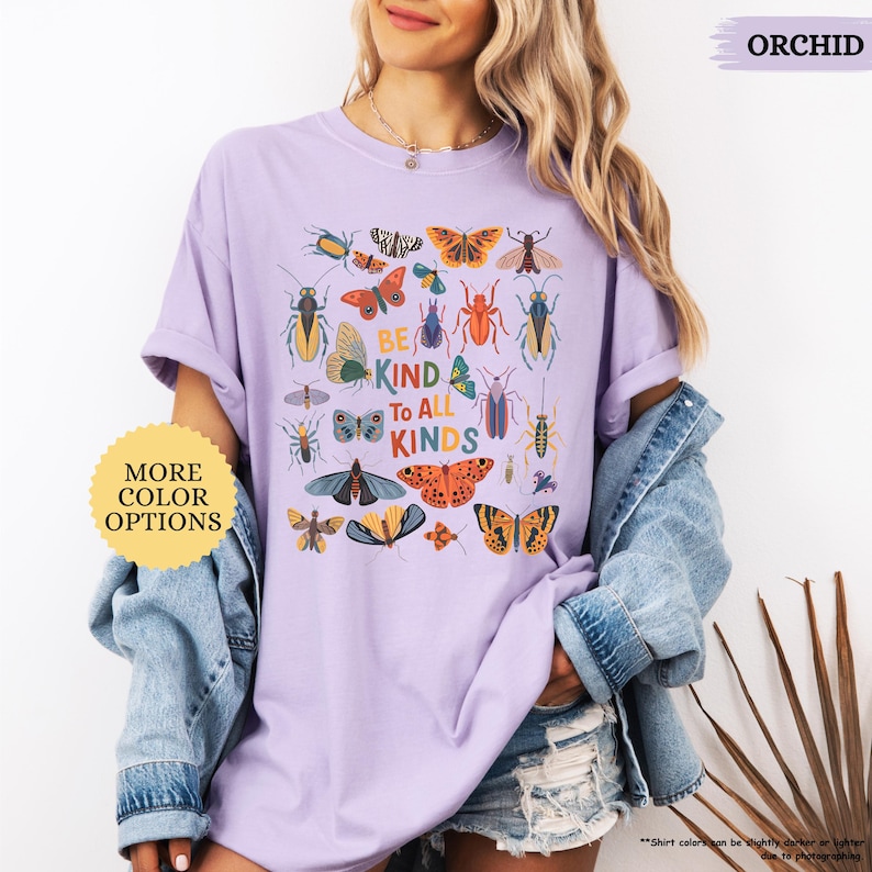 May include: A lilac t-shirt featuring a colourful insect graphic and the words "BE KIND TO ALL KINDS". The shirt is worn with a denim jacket. The text "MORE COLOR OPTIONS" is also visible. The shirt is a light purple colour.