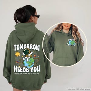 May include: Military green hoodie featuring the text "TOMORROW NEEDS YOU" and an astronaut graphic. The back of the hoodie has a large design with an astronaut on a planet. The front has a smaller graphic.