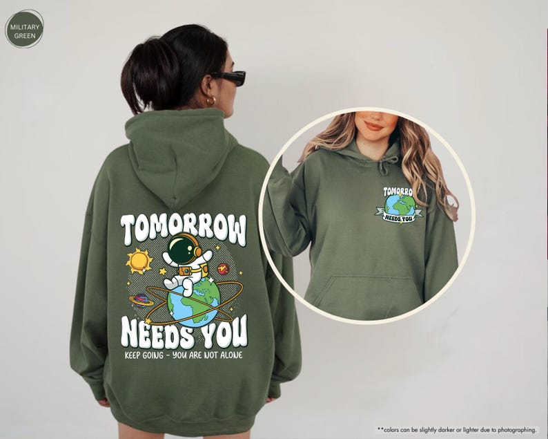 May include: Military green hoodie featuring the text "TOMORROW NEEDS YOU" and an astronaut graphic. The back of the hoodie has a large design with an astronaut on a planet. The front has a smaller graphic.
