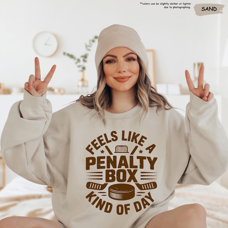 May include: Sand-colored sweatshirt with the text "FEELS LIKE A PENALTY BOX KIND OF DAY" in brown. The design includes hockey sticks, a net, and a puck. The model is wearing a matching beanie.