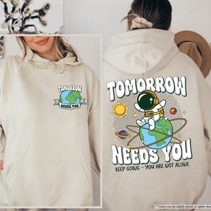May include: Sand-coloured hoodie featuring the text "TOMORROW NEEDS YOU" and an astronaut graphic on Earth. The back displays a larger design with the same text, a sun, planets, and the phrase "KEEP GOING - YOU ARE NOT ALONE."