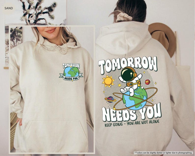 May include: Sand-coloured hoodie featuring the text "TOMORROW NEEDS YOU" and an astronaut graphic on Earth. The back displays a larger design with the same text, a sun, planets, and the phrase "KEEP GOING - YOU ARE NOT ALONE."