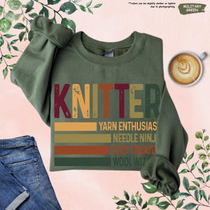 May include: Military green sweatshirt with the word "KNITTER" in a retro font. Below the word are the phrases "YARN ENTHUSIAS", "NEEDLE NINJ", "COZY CREATO", and "WOOL WIZ" in a colorful, striped design.