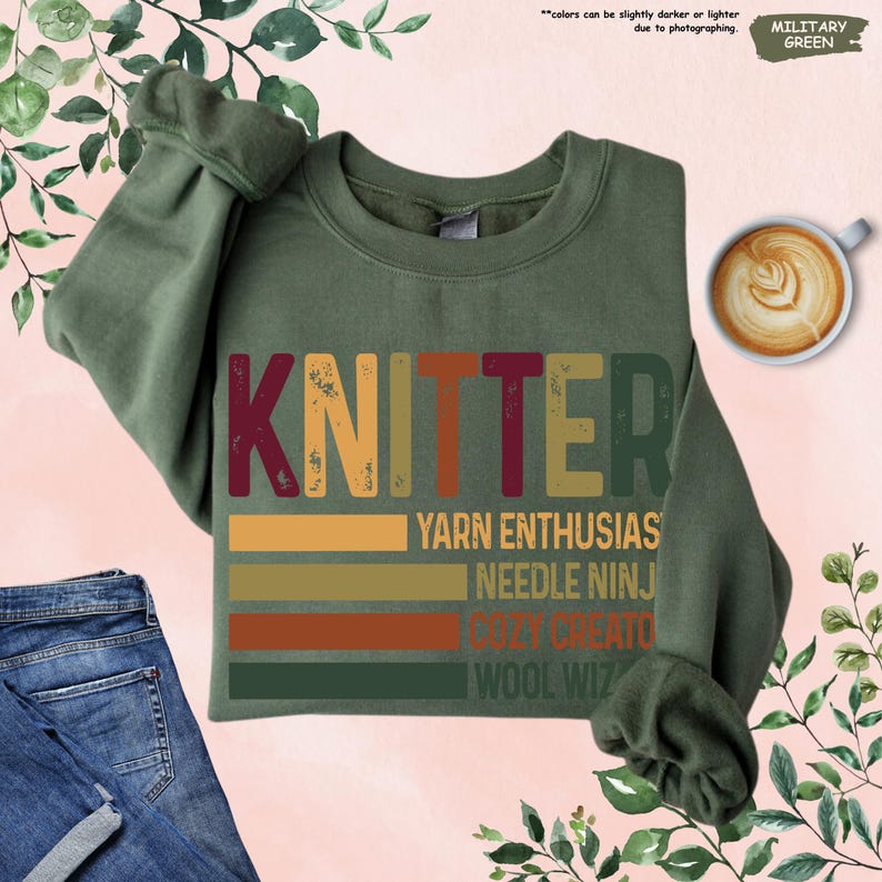 May include: Military green sweatshirt with the word "KNITTER" in a retro font. Below the word are the phrases "YARN ENTHUSIAS", "NEEDLE NINJ", "COZY CREATO", and "WOOL WIZ" in a colorful, striped design.