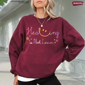 K&ouml;nnte beinhalten: Ein weinroter Pullover mit dem Text "Healing is Not Linear" in Regenbogenfarben mit einem Smiley im Wort "Healing".