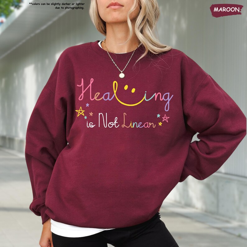 K&ouml;nnte beinhalten: Ein weinroter Pullover mit dem Text "Healing is Not Linear" in Regenbogenfarben mit einem Smiley im Wort "Healing".