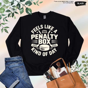 May include: Black sweatshirt with the text "FEELS LIKE A PENALTY BOX KIND OF DAY" in white. The design includes hockey sticks, a puck, and a net. The sweatshirt is paired with blue jeans and a brown tote bag.