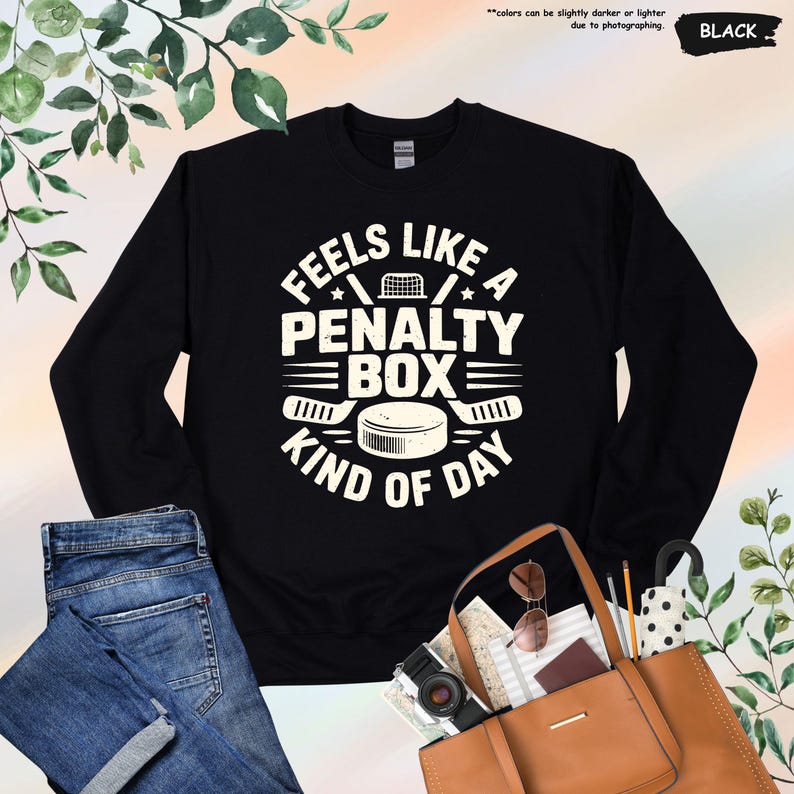 May include: Black sweatshirt with the text "FEELS LIKE A PENALTY BOX KIND OF DAY" in white. The design includes hockey sticks, a puck, and a net. The sweatshirt is paired with blue jeans and a brown tote bag.