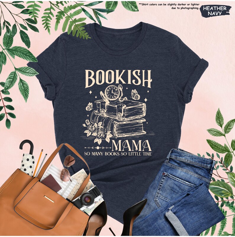 May include: Heather navy t-shirt with the text "BOOKISH MAMA" above a stack of books, a clock, and butterflies. The phrase "SO MANY BOOKS SO LITTLE TIME" is below the graphic. The shirt is paired with blue jeans.