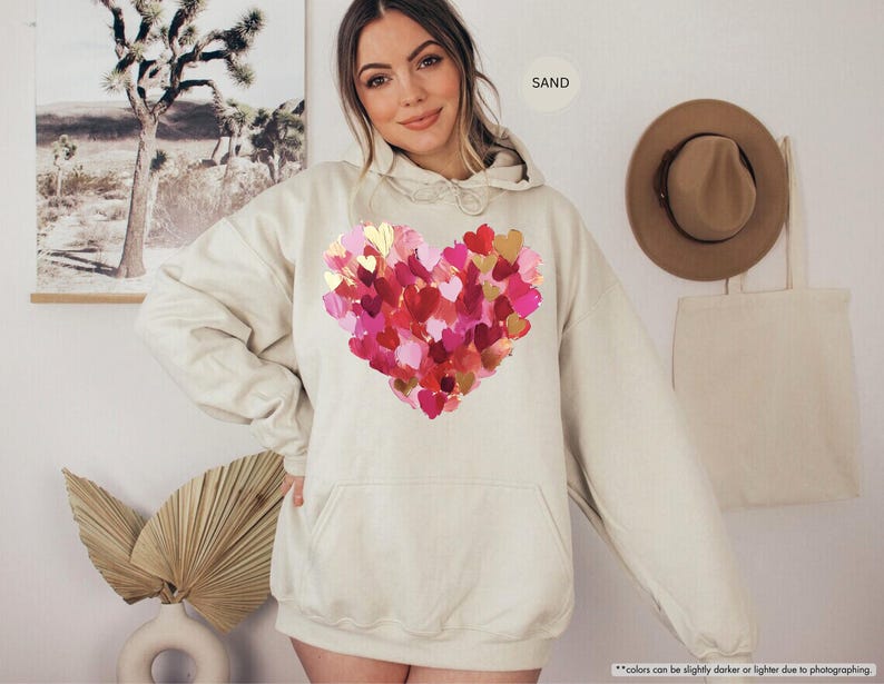 Pu&ograve; includere: Una felpa con cappuccio color sabbia con un grande disegno a cuore composto da pennellate rosa, rosse e dorate sovrapposte. La felpa con cappuccio ha un cappuccio con coulisse e una tasca frontale. La parola "SAND" &egrave; stampata in alto a destra.