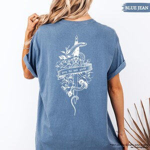 May include: A blue jean-coloured t-shirt with a white graphic on the back. The graphic shows a snake coiled around a sword, flowers, and the text "you do not yield" on a banner. The shirt has rolled-up sleeves.