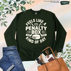 May include: Forest green sweatshirt with the text "Feels Like a Penalty Box Kind of Day" in white. The design includes a hockey net, stars, hockey sticks, and a hockey puck. The sweatshirt is a long-sleeved crew neck.