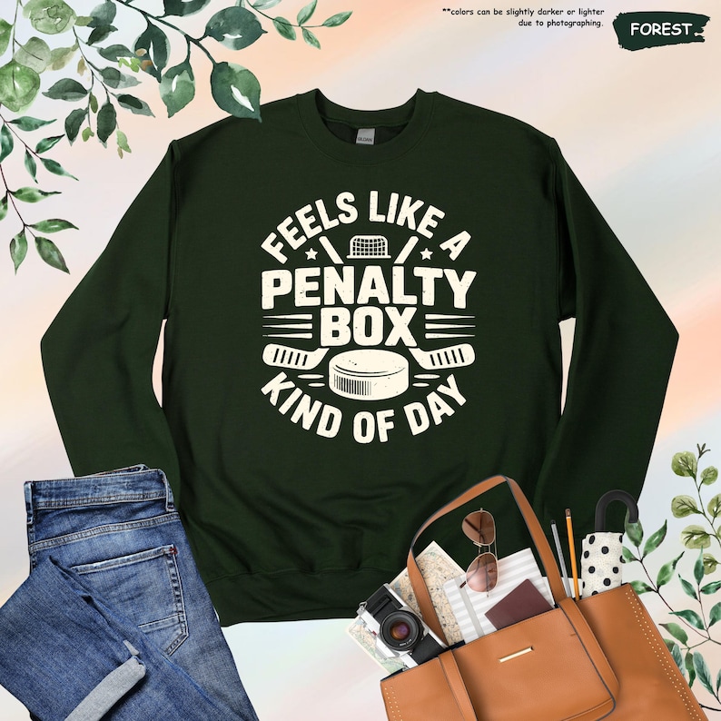 May include: Forest green sweatshirt with the text "Feels Like a Penalty Box Kind of Day" in white. The design includes a hockey net, stars, hockey sticks, and a hockey puck. The sweatshirt is a long-sleeved crew neck.