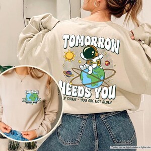 May include: Beige sweatshirt featuring the slogan "TOMORROW NEEDS YOU" with an astronaut graphic. The design includes a sun, planets, and the Earth. A smaller version of the design is on the front.