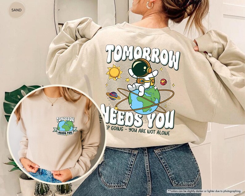 May include: Beige sweatshirt featuring the slogan "TOMORROW NEEDS YOU" with an astronaut graphic. The design includes a sun, planets, and the Earth. A smaller version of the design is on the front.