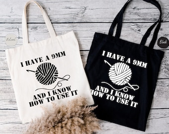 May include: Two tote bags with the text "I have a 9mm and I know how to use it" and a graphic of a ball of yarn and knitting needles. One bag is white and the other is black.