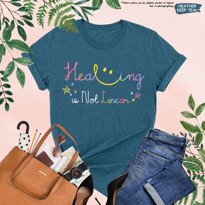 K&ouml;nnte beinhalten: Ein blaues T-Shirt mit dem Text "Healing is Not Linear" in Regenbogenfarben. Der Text enth&auml;lt ein Smiley.
