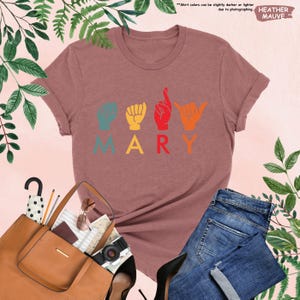 Puede incluir: Una camiseta de color malva con la palabra "MARY" escrita en letras de colores. Cada letra est&aacute; representada por un signo de la lengua de se&ntilde;as americana.