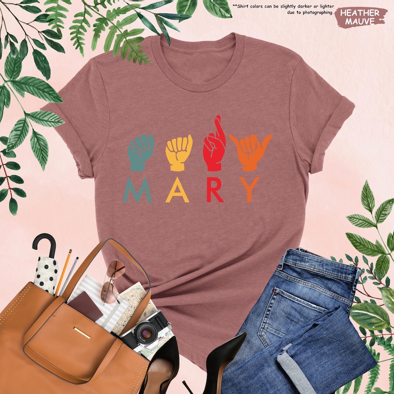 Puede incluir: Una camiseta de color malva con la palabra "MARY" escrita en letras de colores. Cada letra est&aacute; representada por un signo de la lengua de se&ntilde;as americana.
