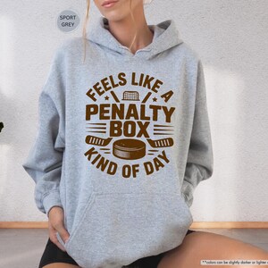 May include: A light grey hoodie with a brown graphic that reads "Feels Like a Penalty Box Kind of Day." The design includes hockey sticks, a puck, and a net. The hoodie has a drawstring hood and a front pocket.