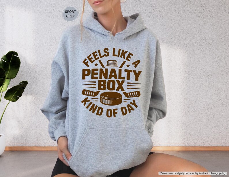 May include: A light grey hoodie with a brown graphic that reads "Feels Like a Penalty Box Kind of Day." The design includes hockey sticks, a puck, and a net. The hoodie has a drawstring hood and a front pocket.