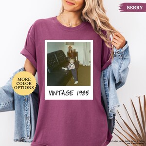 Op de afbeelding: Een maronkleurig T-shirt met een vintage foto van een jong kind dat op een bank zit. De foto is omlijst met een witte rand en heeft de tekst "Vintage 1985" eronder gedrukt.