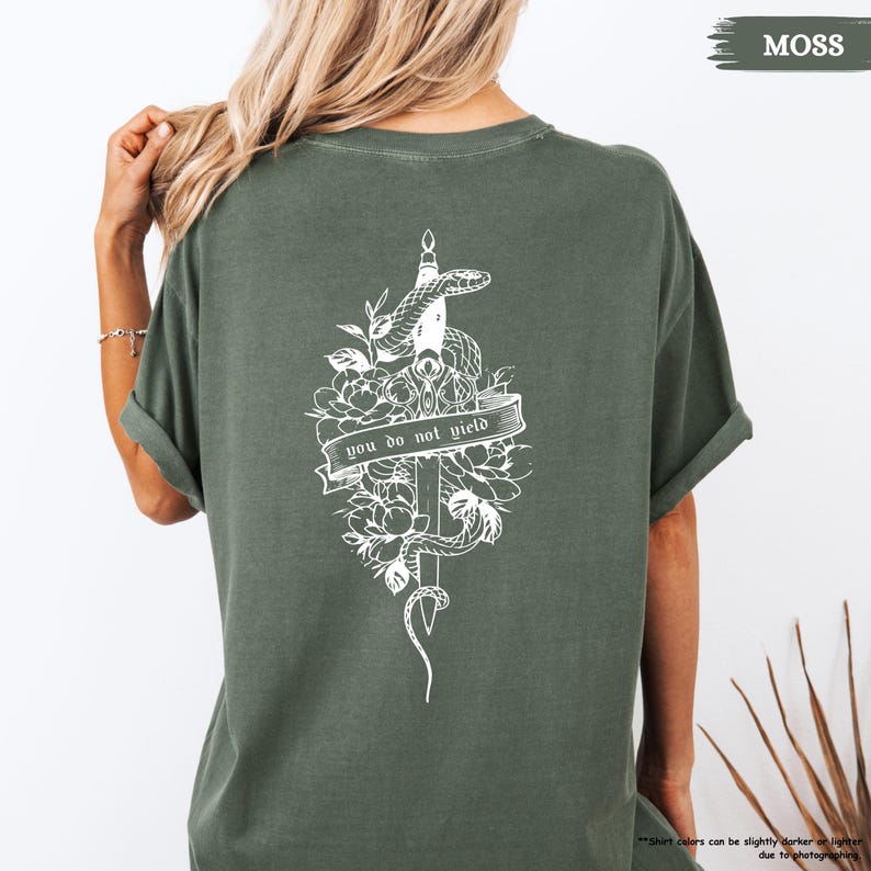 May include: A moss green t-shirt with a white graphic on the back. The graphic shows a snake coiled around a sword, flowers, and the text "YOU DO NOT YIELD" on a banner. The shirt has short sleeves.