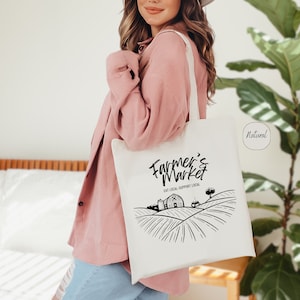 May include: A white tote bag with black text that reads "Farmer's Market" and "Eat Local, Support Local". The bag also features a black and white illustration of a farm scene with a barn, tractor, and fields.