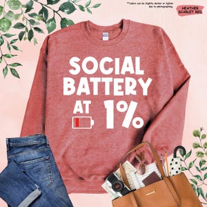 May include: Heather scarlet red sweatshirt with the text "SOCIAL BATTERY AT 1%" in white. The design includes a low battery icon. The sweatshirt is paired with blue jeans and a brown tote bag.