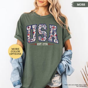 May include: A green t-shirt with a floral print of the word "USA" and the text "EST. 1776".