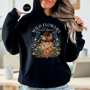 May include: Black hoodie with a white graphic print of a horseshoe with flowers and a woman wearing a hat. The text on the graphic reads "WILD FLOWERS wild horses".