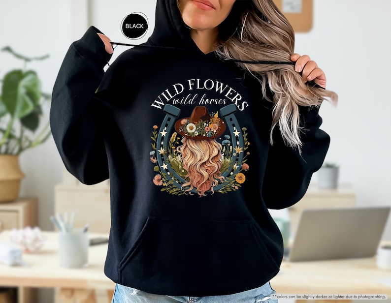 May include: Black hoodie with a white graphic print of a horseshoe with flowers and a woman wearing a hat. The text on the graphic reads "WILD FLOWERS wild horses".