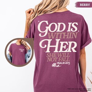 May include: Berry-colored t-shirt with the text "God is Within Her, She Will Not Fall, Psalm 46:5" in a white and tan font. The shirt has a crew neck and short sleeves. A smaller image shows the front of the shirt with "Psalm 465" printed on the chest.