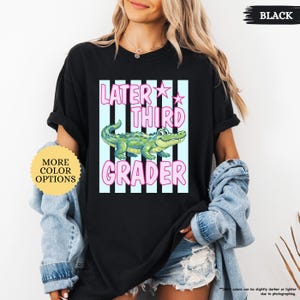 May include: Black t-shirt with the text "LATER THIRD GRADER" in pink, with a cartoon alligator illustration. The shirt also has a "MORE COLOR OPTIONS" button.