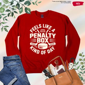 May include: Red sweatshirt with the text "Feels Like A Penalty Box Kind Of Day" in white. The design includes hockey sticks, a puck, and a net. The word "RED" is in the upper right corner. The sweatshirt is paired with blue jeans and a brown tote bag.