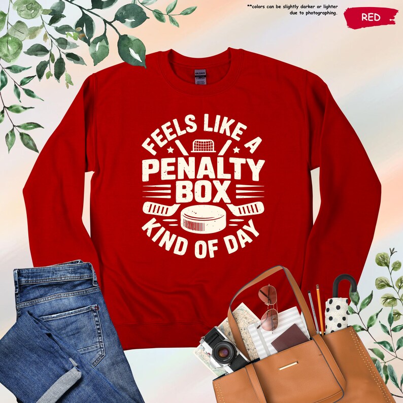 May include: Red sweatshirt with the text "Feels Like A Penalty Box Kind Of Day" in white. The design includes hockey sticks, a puck, and a net. The word "RED" is in the upper right corner. The sweatshirt is paired with blue jeans and a brown tote bag.
