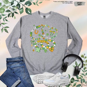 K&ouml;nnte beinhalten: Ein graues Sweatshirt mit einem gr&uuml;nen, gelben und wei&szlig;en Design aus drei Halsketten mit Anh&auml;ngern. Die Halsketten tragen die Worte "One Lucky Charm".