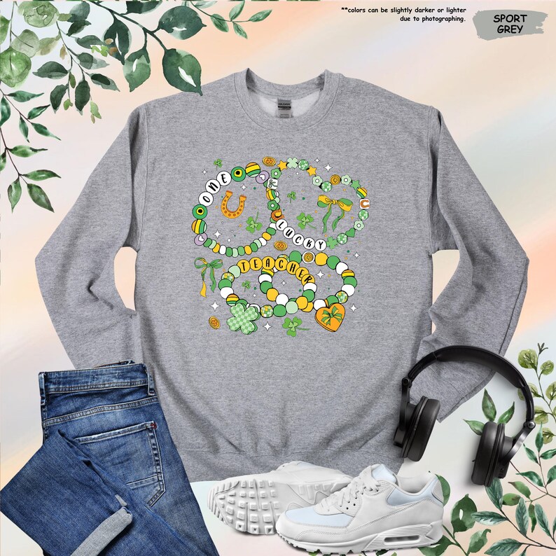 K&ouml;nnte beinhalten: Ein graues Sweatshirt mit einem gr&uuml;nen, gelben und wei&szlig;en Design aus drei Halsketten mit Anh&auml;ngern. Die Halsketten tragen die Worte "One Lucky Charm".