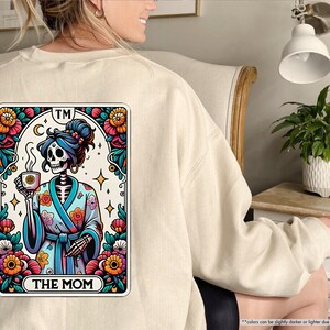 May include: A beige sweatshirt with a colorful illustration of a skeleton wearing a floral robe and holding a cup of coffee. The illustration is framed in a card with the text "TM THE MOM" at the bottom.