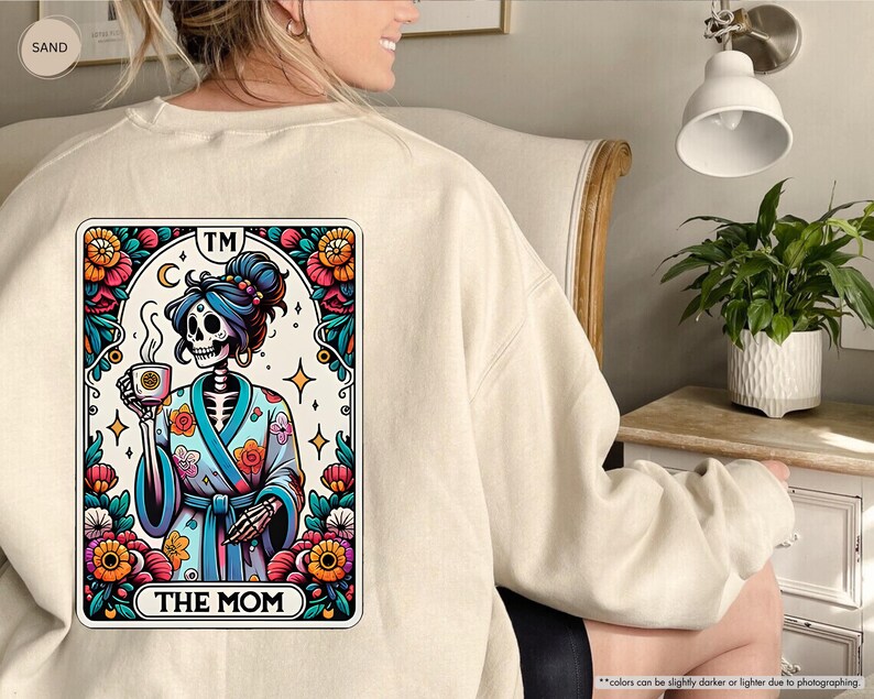 May include: A beige sweatshirt with a colorful illustration of a skeleton wearing a floral robe and holding a cup of coffee. The illustration is framed in a card with the text "TM THE MOM" at the bottom.