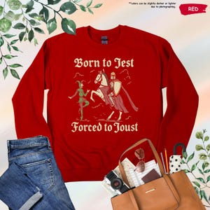 K&ouml;nnte beinhalten: Ein rotes Sweatshirt mit dem Text "Born to Jest Forced to Joust" in einer mittelalterlichen Schriftart. Die Grafik zeigt einen Narren und einen Ritter zu Pferd. Das Sweatshirt wird mit einer Jeans und einer braunen Tragetasche pr&auml;sentiert.