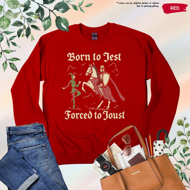 K&ouml;nnte beinhalten: Ein rotes Sweatshirt mit dem Text "Born to Jest Forced to Joust" in einer mittelalterlichen Schriftart. Die Grafik zeigt einen Narren und einen Ritter zu Pferd. Das Sweatshirt wird mit einer Jeans und einer braunen Tragetasche pr&auml;sentiert.
