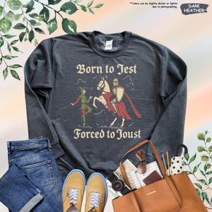 K&ouml;nnte beinhalten: Dunkelgraues Sweatshirt mit dem Text "Born to Jest Forced to Joust" und einer Grafik eines Narren und eines Ritters zu Pferd. Das Sweatshirt wird mit Jeans, Schuhen und einer Handtasche pr&auml;sentiert.