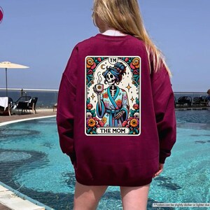 May include: A maroon sweatshirt with a colorful graphic of a skeleton wearing a robe and holding a cup. The graphic is in the style of a tarot card and says "TM THE MOM" at the bottom.