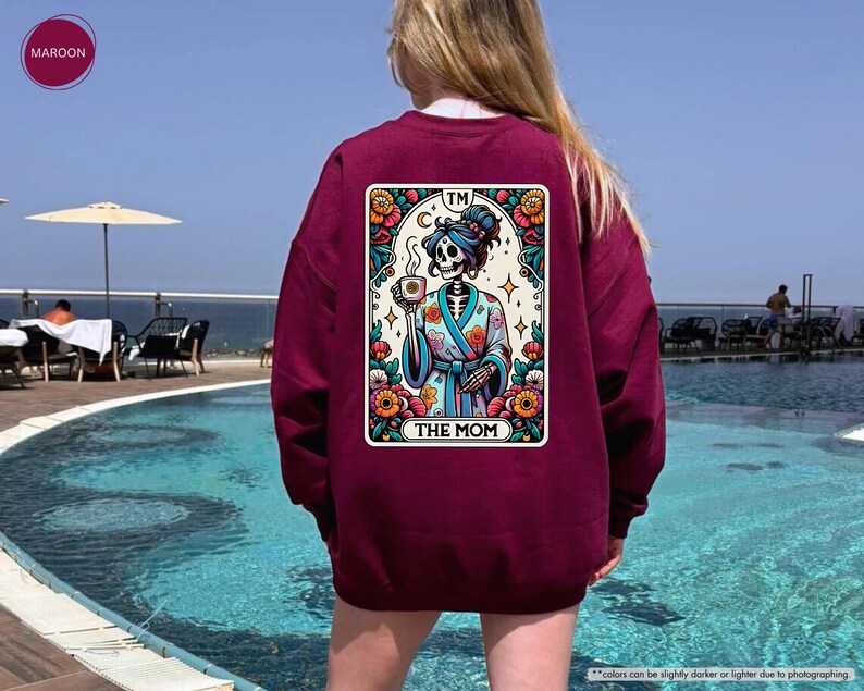 May include: A maroon sweatshirt with a colorful graphic of a skeleton wearing a robe and holding a cup. The graphic is in the style of a tarot card and says "TM THE MOM" at the bottom.