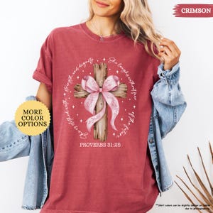Op de afbeelding: Een roodbruin t-shirt met een verweerde look heeft een houten kruis met een roze strik. Het kruis is omringd door een cirkel van witte sterren en de tekst "Zij lacht zonder angst voor de toekomst" en "Zij is bekleed met kracht en waardigheid". De onderkant van het ontwerp bevat de tekst "Spreuken 31:25".
